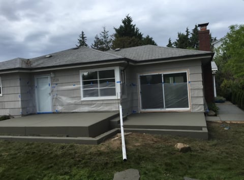 Single-story residential house with gray siding, covered porch, and evergreen trees in background under cloudy sky