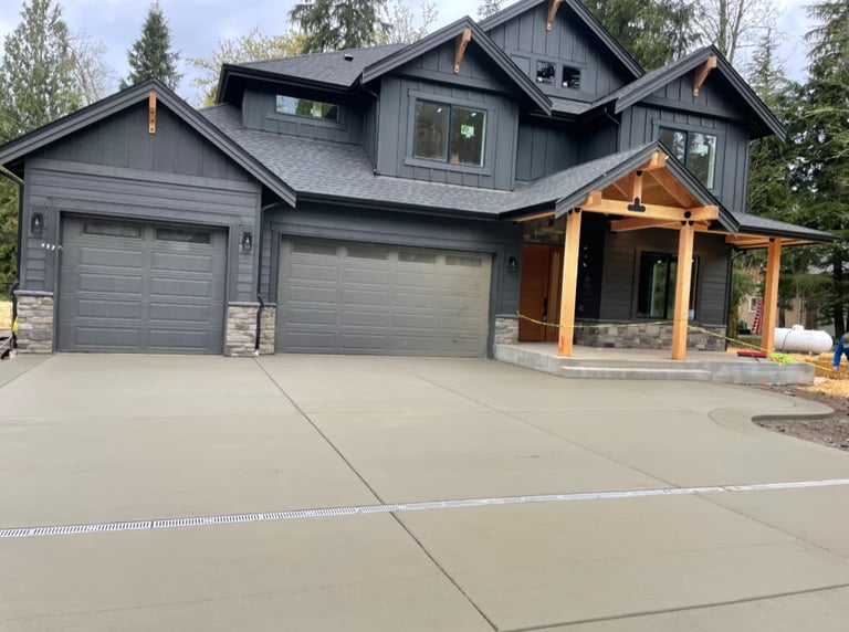 Modern two-story dark gray house with three-car garage, wooden front porch, and concrete driveway surrounded by evergreen trees