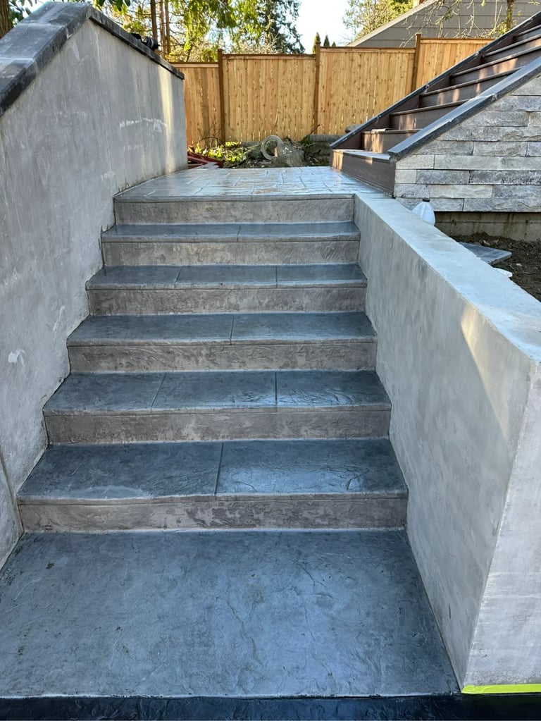 Concrete outdoor staircase with metal handrail leading upward between white walls toward a wooden fence and backyard patio area