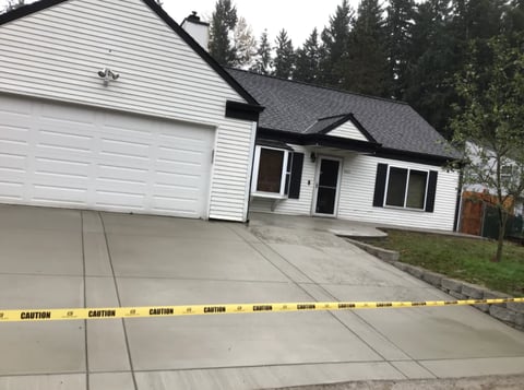 White house with attached garage and driveway marked with yellow caution tape, surrounded by evergreen trees
