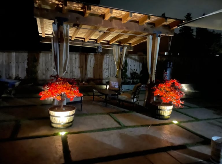 Nighttime outdoor patio with wooden pergola, illuminated red flowering trees in planters, string lights, and lounge seating