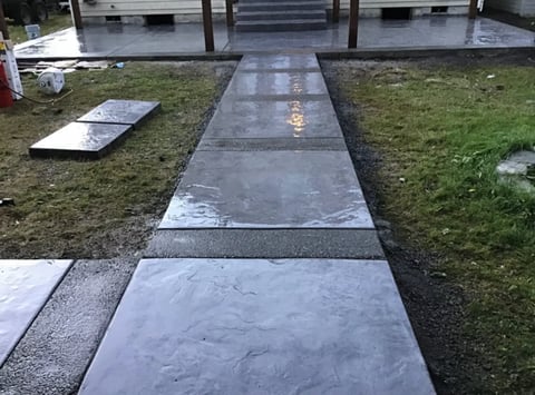 Concrete pathway with grave markers in a cemetery, lined with grass on both sides and buildings in the background
