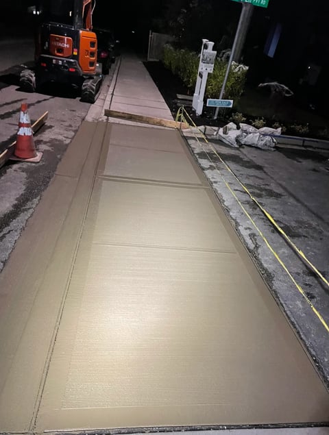 Newly poured concrete sidewalk at night with construction equipment and safety barriers visible
