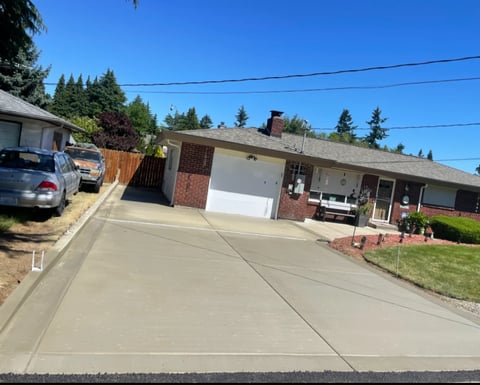 Suburban residential street with single-story brick house, concrete driveway, parked car, and evergreen trees under clear blue sky