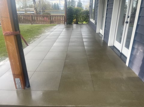 Covered patio with gray concrete flooring extending from house toward backyard with lawn and wooden fence visible