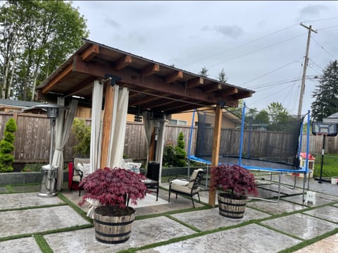 Wooden pergola with seating area, decorative red maple plants in barrel planters, trampoline and residential backyard setting
